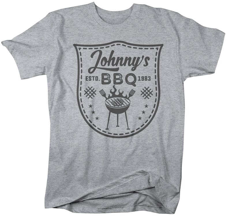 May include: A gray t-shirt featuring a shield-shaped graphic with the text "Johnny's BBQ ESTD. 1983." The graphic includes a grill, flames, and stars. The shirt is a casual wear item.