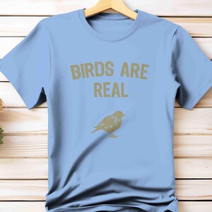 K&ouml;nnte beinhalten: Hellblaues T-Shirt mit dem Text "BIRDS ARE REAL" in goldfarbenen Buchstaben. Unter dem Text befindet sich eine goldfarbene Vogelgrafik. Das T-Shirt h&auml;ngt an einem Holzb&uuml;gel vor einem hellen Holz Hintergrund.