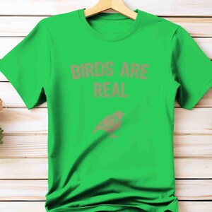 K&ouml;nnte beinhalten: Ein leuchtend gr&uuml;nes T-Shirt mit dem Text "BIRDS ARE REAL" in Hellbraun und einer passenden Vogelgrafik. Das Shirt wird vor einem Holzuntergrund pr&auml;sentiert.