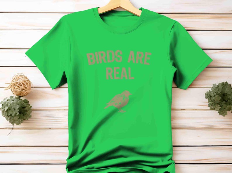 K&ouml;nnte beinhalten: Ein leuchtend gr&uuml;nes T-Shirt mit dem Text "BIRDS ARE REAL" in Hellbraun und einer passenden Vogelgrafik. Das Shirt wird vor einem Holzuntergrund pr&auml;sentiert.