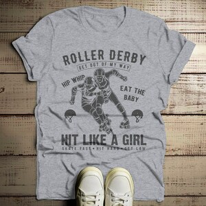 May include: Gray t-shirt with a roller derby graphic. The graphic features two roller derby players skating and the text "Roller Derby Get Out Of My Way Hip Whip Eat The Baby Hit Like A Girl Skate Fast Hit Hard Get Low".