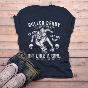 May include: A navy blue t-shirt with a white graphic of two roller derby players. The text on the shirt reads "Roller Derby Get Out of My Way Hip Whip Eat the Baby Hit Like a Girl Skate Fast Hit Hard Get Low".