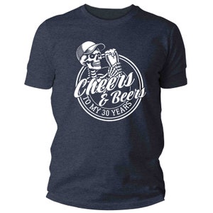 May include: Navy blue t-shirt with a white graphic of a skeleton in a cap, toasting with a beer, and the text "Cheers & Beers To My 30 Years" in a circle. The shirt is a crew neck style.