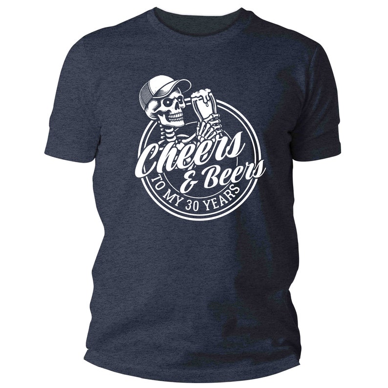 May include: Navy blue t-shirt with a white graphic of a skeleton in a cap, toasting with a beer, and the text "Cheers & Beers To My 30 Years" in a circle. The shirt is a crew neck style.