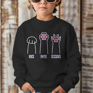 May include: Black sweatshirt featuring a white graphic of cat paws representing rock, paper, and scissors. The words "ROCK", "PAPER", and "SCISSORS" are printed below the corresponding paw images. The sweatshirt is worn by a person wearing sunglasses.