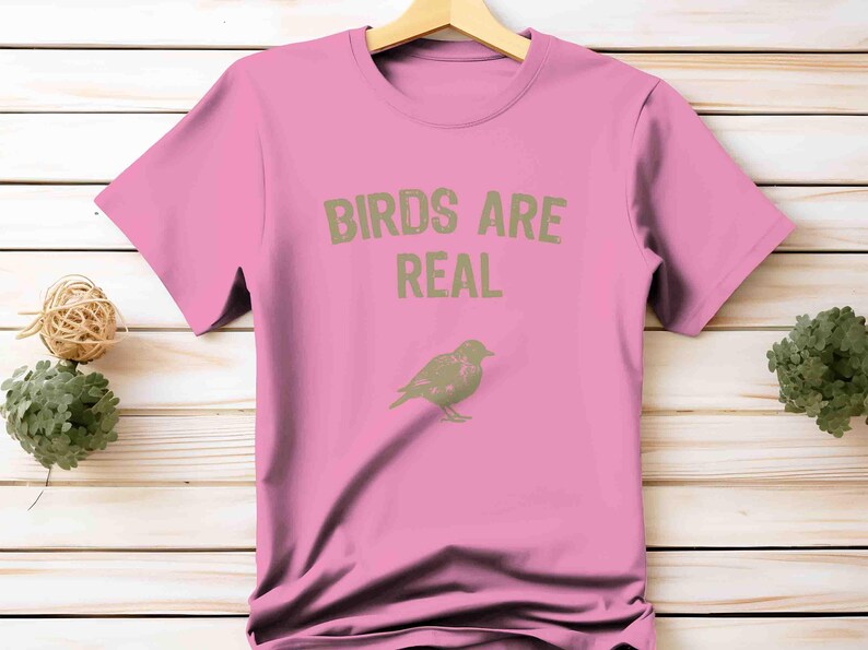 K&ouml;nnte beinhalten: Ein pinkfarbenes T-Shirt mit dem Text "BIRDS ARE REAL" in Hellgr&uuml;n. Darunter befindet sich eine hellgr&uuml;ne Illustration eines Vogels. Das T-Shirt h&auml;ngt an einem Holzb&uuml;gel.