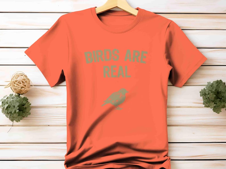 K&ouml;nnte beinhalten: Ein orangefarbenes T-Shirt mit dem Text "BIRDS ARE REAL" in gr&uuml;nen Buchstaben und einer gr&uuml;nen Vogelgrafik. Das T-Shirt h&auml;ngt an einem Holzb&uuml;gel.