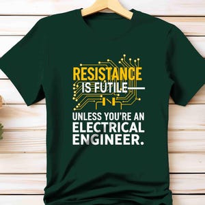 May include: Dark green t-shirt with the text "RESISTANCE IS FUTILE UNLESS YOU'RE AN ELECTRICAL ENGINEER." The text is in white and yellow, with a circuit board graphic. The shirt is hanging on a wooden hanger.