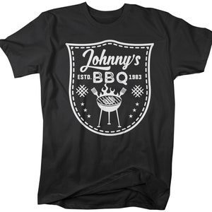 May include: Black t-shirt with a white graphic. The graphic is a shield shape with the words "Johnny's BBQ ESTD. 1983" and a grill illustration. The design includes stars, hash marks, and a dotted border.