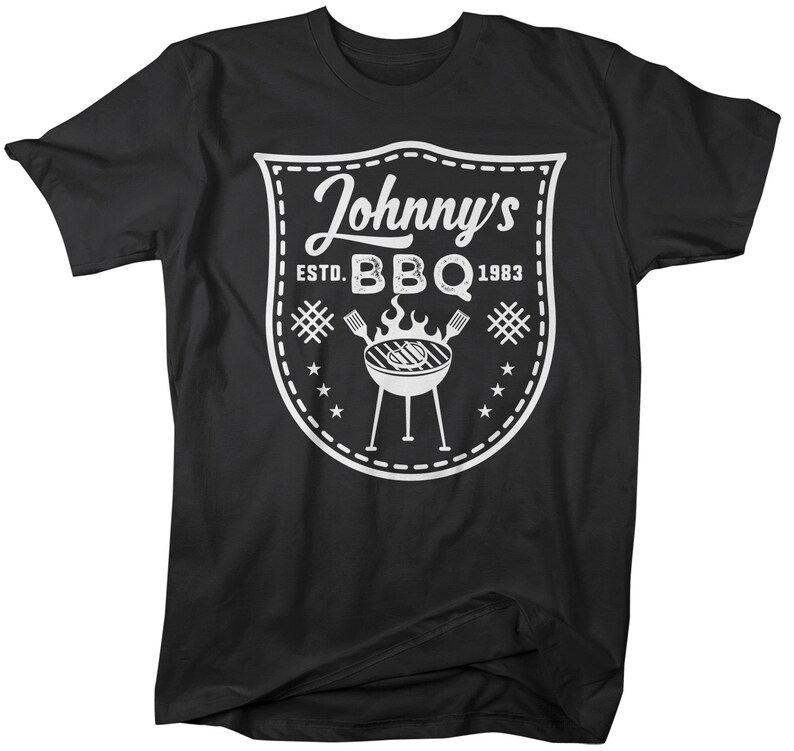 May include: Black t-shirt with a white graphic. The graphic is a shield shape with the words "Johnny's BBQ ESTD. 1983" and a grill illustration. The design includes stars, hash marks, and a dotted border.