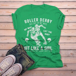 May include: Green t-shirt with a white graphic of two roller derby players. The text reads "Roller Derby Get Out of My Way Hip Whip Eat the Baby Hit Like a Girl Skate Fast Hit Hard Get Low".