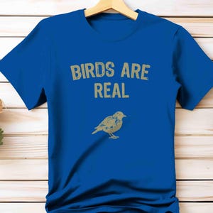 K&ouml;nnte beinhalten: Ein k&ouml;nigsblaues T-Shirt mit dem Text "BIRDS ARE REAL" in einer verblassten, hellbraunen Schrift. Darunter befindet sich eine hellbraune Illustration eines Vogels. Das T-Shirt h&auml;ngt an einem Holzkleiderb&uuml;gel.