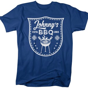 May include: A blue t-shirt featuring a white graphic. The graphic is a shield shape with the words "Johnny's BBQ ESTD. 1983" and a grill illustration. The shirt is a short-sleeved crew neck.