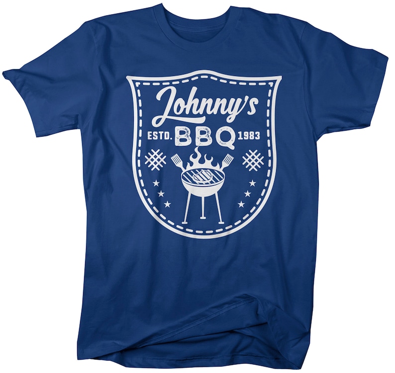 May include: A blue t-shirt featuring a white graphic. The graphic is a shield shape with the words "Johnny's BBQ ESTD. 1983" and a grill illustration. The shirt is a short-sleeved crew neck.