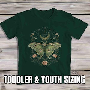 May include: Dark green t-shirt featuring a detailed illustration of a moth with a crescent moon, flowers, and celestial elements. The text "TODDLER & YOUTH SIZING" is displayed at the bottom. The design is centered on the shirt.