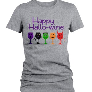 K&ouml;nnte beinhalten: Graues T-Shirt mit dem Text "Happy Hallo-wine" in Lila. F&uuml;nf Weingl&auml;ser mit verschiedenen Halloween-Charaktergesichtern sind unter dem Text aufgereiht.