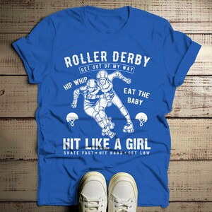 May include: Blue t-shirt with a white graphic of two roller derby skaters. The text on the shirt reads "Roller Derby Get Out Of My Way Hip Whip Eat The Baby Hit Like A Girl Skate Fast Hit Hard Get Low".