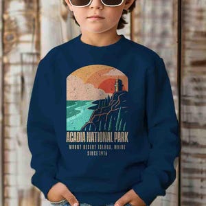 May include: A navy blue sweatshirt with a vintage-style graphic of a lighthouse on a cliff overlooking the ocean. The graphic includes the text "Acadia National Park, Mount Desert Island, Maine Since 1916".