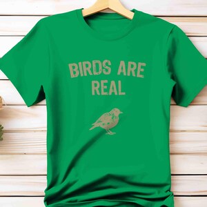 K&ouml;nnte beinhalten: Ein gr&uuml;nes T-Shirt mit dem Text "BIRDS ARE REAL" in Hellbraun und einer Vogelillustration darunter. Das T-Shirt h&auml;ngt an einem Holzb&uuml;gel. Der Hintergrund ist eine Holzoberfl&auml;che.