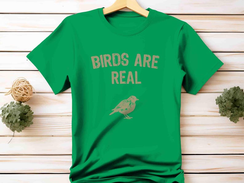 K&ouml;nnte beinhalten: Ein gr&uuml;nes T-Shirt mit dem Text "BIRDS ARE REAL" in Hellbraun und einer Vogelillustration darunter. Das T-Shirt h&auml;ngt an einem Holzb&uuml;gel. Der Hintergrund ist eine Holzoberfl&auml;che.