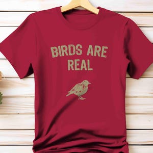 K&ouml;nnte beinhalten: Ein rotes T-Shirt mit dem Text "BIRDS ARE REAL" in Hellbraun und einer Vogelillustration. Das Shirt h&auml;ngt an einem Holzb&uuml;gel vor einem hellen Holz Hintergrund.