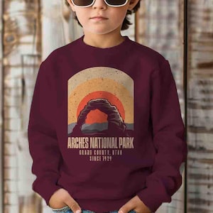 May include: A maroon sweatshirt with a vintage-style graphic of Delicate Arch in Arches National Park, Utah. The graphic features a sunset with a red sun and the text "Arches National Park, Grand County, Utah, Since 1929."
