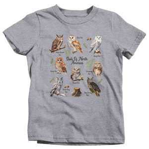 May include: A gray t-shirt with a graphic of different types of owls from North America. The owls are illustrated in a watercolor style and are surrounded by branches and leaves. The text "Owls Of North America" is printed in black at the center of the design.