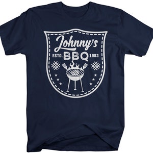 May include: Navy blue t-shirt with a white graphic. The graphic is a shield shape with the text "Johnny's BBQ ESTD. 1983". The design includes a grill, flames, and stars. A casual tee for barbecue enthusiasts.