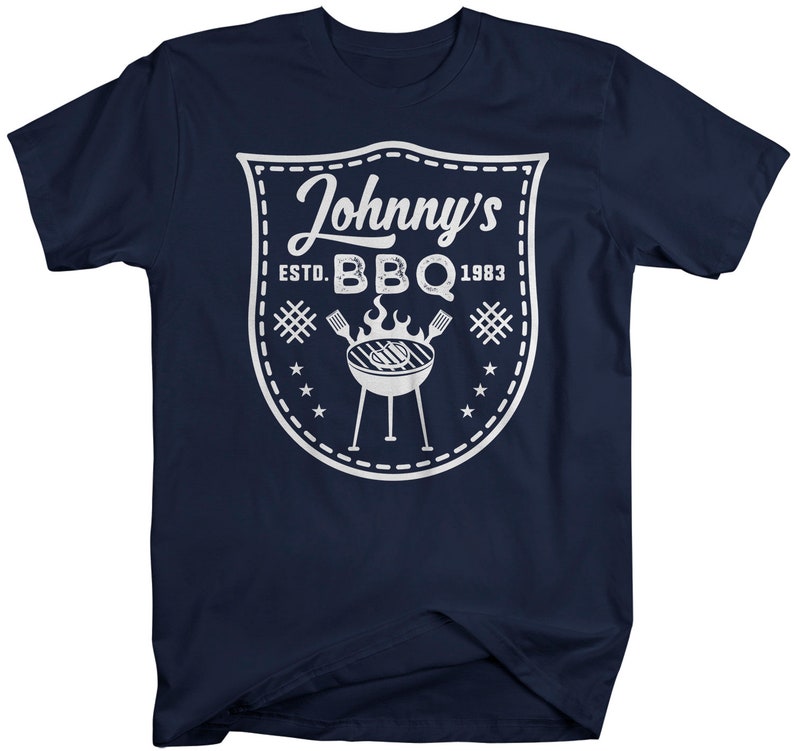 May include: Navy blue t-shirt with a white graphic. The graphic is a shield shape with the text "Johnny's BBQ ESTD. 1983". The design includes a grill, flames, and stars. A casual tee for barbecue enthusiasts.