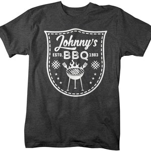 May include: Dark gray t-shirt with a white graphic. The graphic is a shield-shaped design with the words "Johnny's BBQ Estd. 1983". The design includes a grill, flames, stars, and crossed utensils.