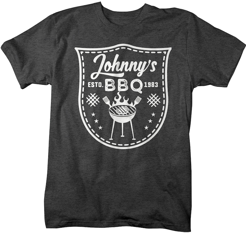 May include: Dark gray t-shirt with a white graphic. The graphic is a shield-shaped design with the words "Johnny's BBQ Estd. 1983". The design includes a grill, flames, stars, and crossed utensils.