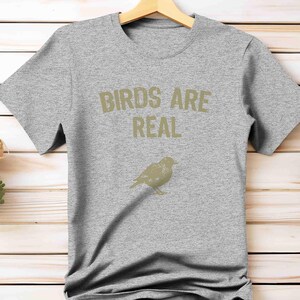 K&ouml;nnte beinhalten: Graues T-Shirt mit dem Text "BIRDS ARE REAL" in hellem Braun und einer Vogelillustration. Das T-Shirt h&auml;ngt an einem Holzb&uuml;gel.