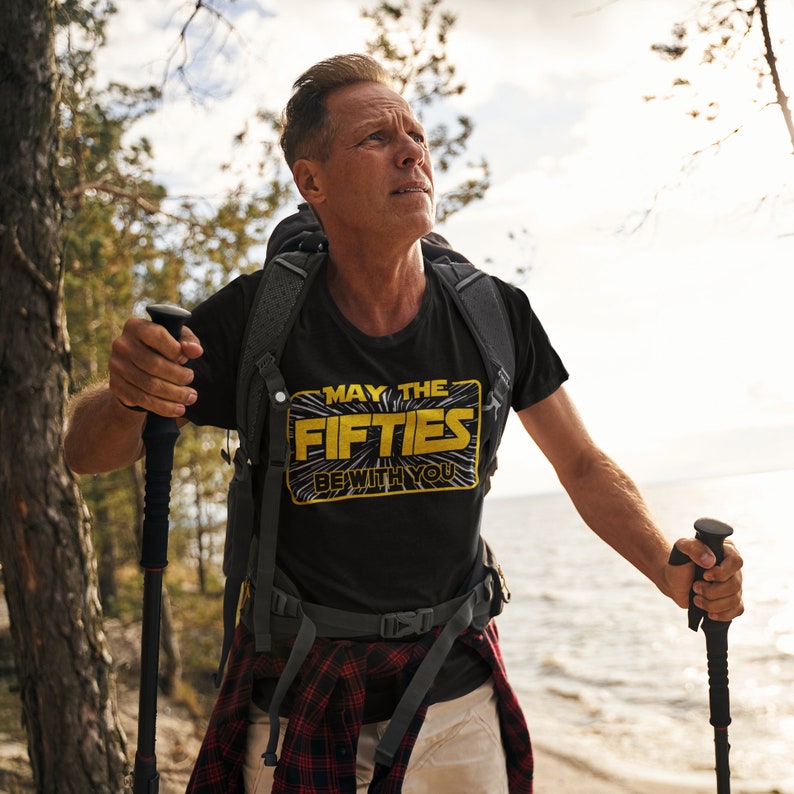 K&ouml;nnte beinhalten: Ein Mann tr&auml;gt ein schwarzes T-Shirt mit dem Text "May the Fifties Be With You" in gelben Buchstaben. Er wandert in einem Wald und tr&auml;gt einen Rucksack und Wanderst&ouml;cke.