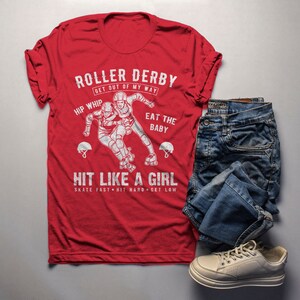 May include: Red t-shirt with a white graphic of two roller derby players. The graphic says "Roller Derby Get Out Of My Way", "Hip Whip", "Eat The Baby", "Hit Like A Girl", and "Skate Fast Hit Hard Get Low".