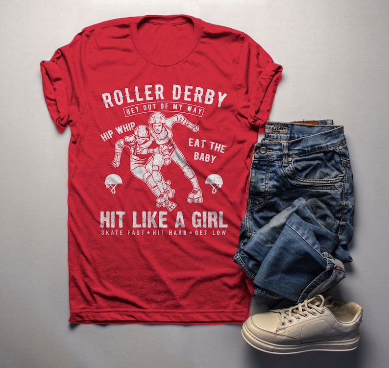 May include: Red t-shirt with a white graphic of two roller derby players. The graphic says "Roller Derby Get Out Of My Way", "Hip Whip", "Eat The Baby", "Hit Like A Girl", and "Skate Fast Hit Hard Get Low".