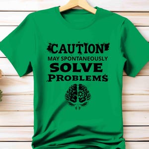 May include: A bright green t-shirt with the text "CAUTION MAY SPONTANEOUSLY SOLVE PROBLEMS" in black. A black brain graphic is below the text. The shirt is hanging on a wooden hanger.