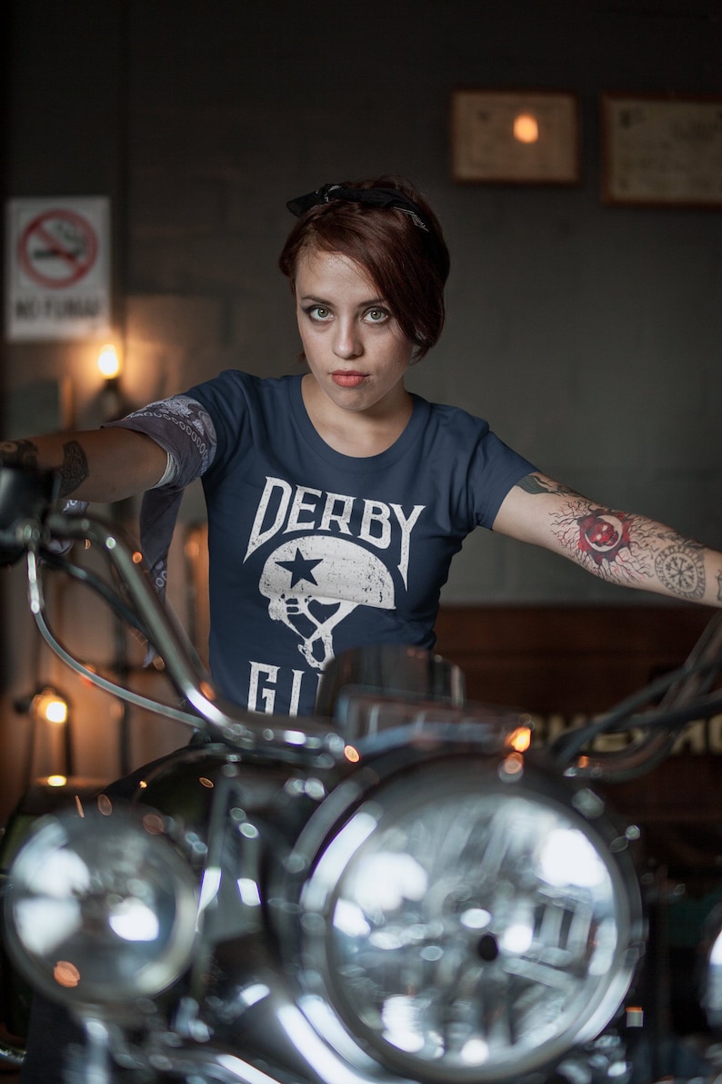 May include: A woman wearing a navy blue t-shirt with the text "Derby Girl" and a white helmet graphic. She is sitting on a motorcycle with a chrome headlight.