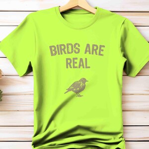 K&ouml;nnte beinhalten: Neongr&uuml;nes T-Shirt mit dem grauen Schriftzug "BIRDS ARE REAL" und einer grauen Vogelillustration. Das T-Shirt h&auml;ngt an einem Holzb&uuml;gel vor einem hellen Holz Hintergrund.