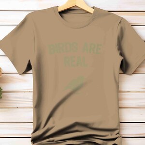 K&ouml;nnte beinhalten: Ein beiges T-Shirt mit dem Text "BIRDS ARE REAL" in gr&uuml;nen Buchstaben. Unter dem Text befindet sich eine gr&uuml;ne Vogelgrafik. Das T-Shirt h&auml;ngt an einem Holzb&uuml;gel vor einem hellen Holzgrund.