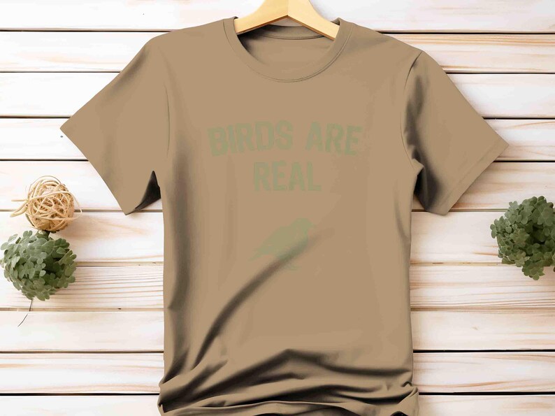 K&ouml;nnte beinhalten: Ein beiges T-Shirt mit dem Text "BIRDS ARE REAL" in gr&uuml;nen Buchstaben. Unter dem Text befindet sich eine gr&uuml;ne Vogelgrafik. Das T-Shirt h&auml;ngt an einem Holzb&uuml;gel vor einem hellen Holzgrund.