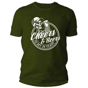 May include: Dark olive green t-shirt with a white graphic. The graphic shows a skeleton in a cap, holding a beer, and the text "Cheers & Beers To My 30 Years" in a circular design. A casual wear item.