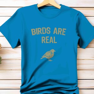 K&ouml;nnte beinhalten: T&uuml;rkisblaues T-Shirt mit dem Text "BIRDS ARE REAL" in verblichener goldener Schrift. Darunter eine goldene Illustration eines Vogels. Das T-Shirt h&auml;ngt an einem Holzkleiderb&uuml;gel.
