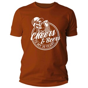 May include: A rust-coloured t-shirt with a white graphic. The graphic shows a skeleton in a cap, holding a beer, with the text "Cheers & Beers To My 30 Years" in a circle. The shirt has short sleeves and a crew neck.