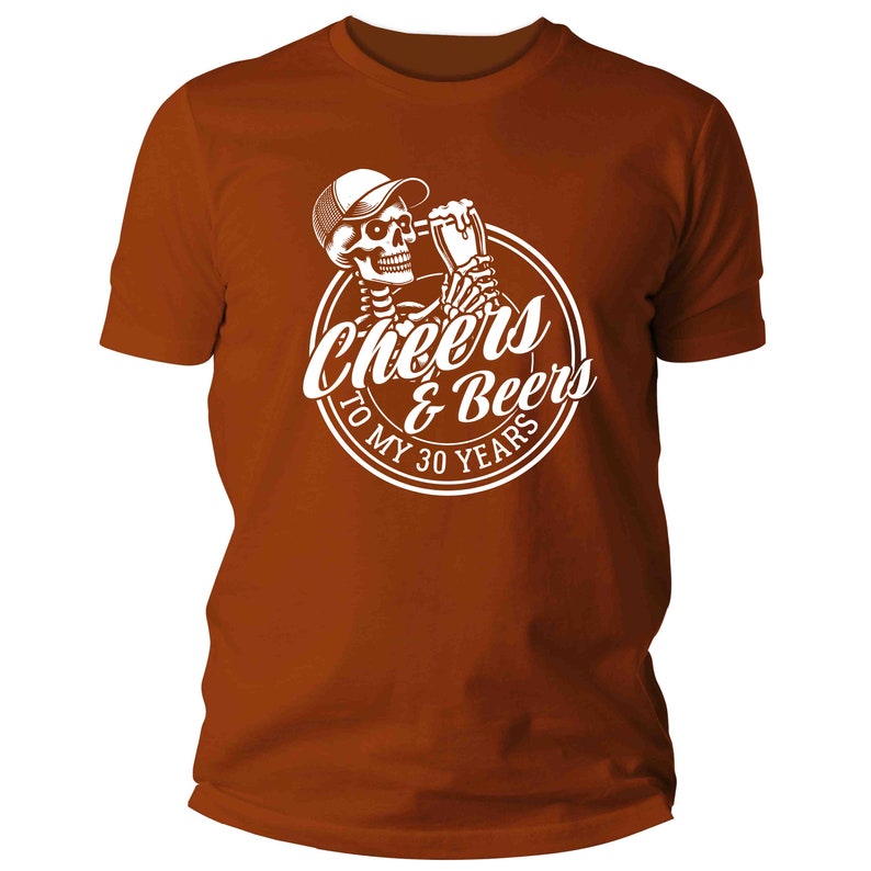 May include: A rust-coloured t-shirt with a white graphic. The graphic shows a skeleton in a cap, holding a beer, with the text "Cheers & Beers To My 30 Years" in a circle. The shirt has short sleeves and a crew neck.