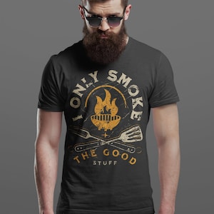May include: A black t-shirt with a vintage-style graphic that says "I Only Smoke The Good Stuff". The graphic features a grill with flames, crossed grilling utensils, and a distressed circle around the text.