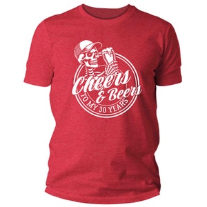 May include: Red t-shirt featuring a white graphic of a skeleton in a cap, toasting with a beer. The graphic reads "Cheers & Beers To My 30 Years" in a circular design. The shirt is made of a soft, textured material.