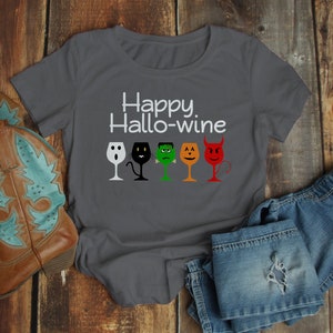 K&ouml;nnte beinhalten: Ein dunkelgraues T-Shirt mit dem Text "Happy Hallo-wine" und einer Reihe von f&uuml;nf Weingl&auml;sern mit Halloween-Charaktergesichtern. Die Gl&auml;ser sind ein Geist, eine schwarze Katze, Frankenstein, ein K&uuml;rbis und ein Teufel.