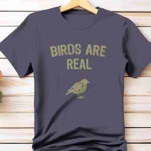 K&ouml;nnte beinhalten: Ein marineblaues T-Shirt mit dem Text "BIRDS ARE REAL" in verblichener, hellbrauner Schrift. Darunter befindet sich eine hellbraune Illustration eines Vogels. Das T-Shirt h&auml;ngt an einem Holzb&uuml;gel.