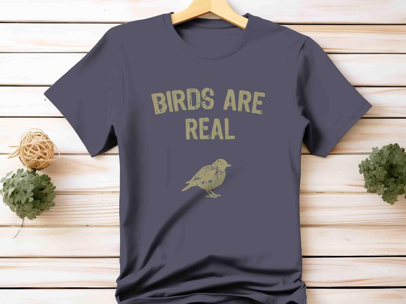 K&ouml;nnte beinhalten: Ein marineblaues T-Shirt mit dem Text "BIRDS ARE REAL" in verblichener, hellbrauner Schrift. Darunter befindet sich eine hellbraune Illustration eines Vogels. Das T-Shirt h&auml;ngt an einem Holzb&uuml;gel.