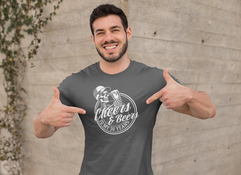 May include: Dark grey t-shirt with a white graphic. The graphic shows a skeleton wearing a baseball cap and holding a beer, with the words "Cheers & Beers To My 30 Years" in a circular design. The t-shirt is being pointed at.